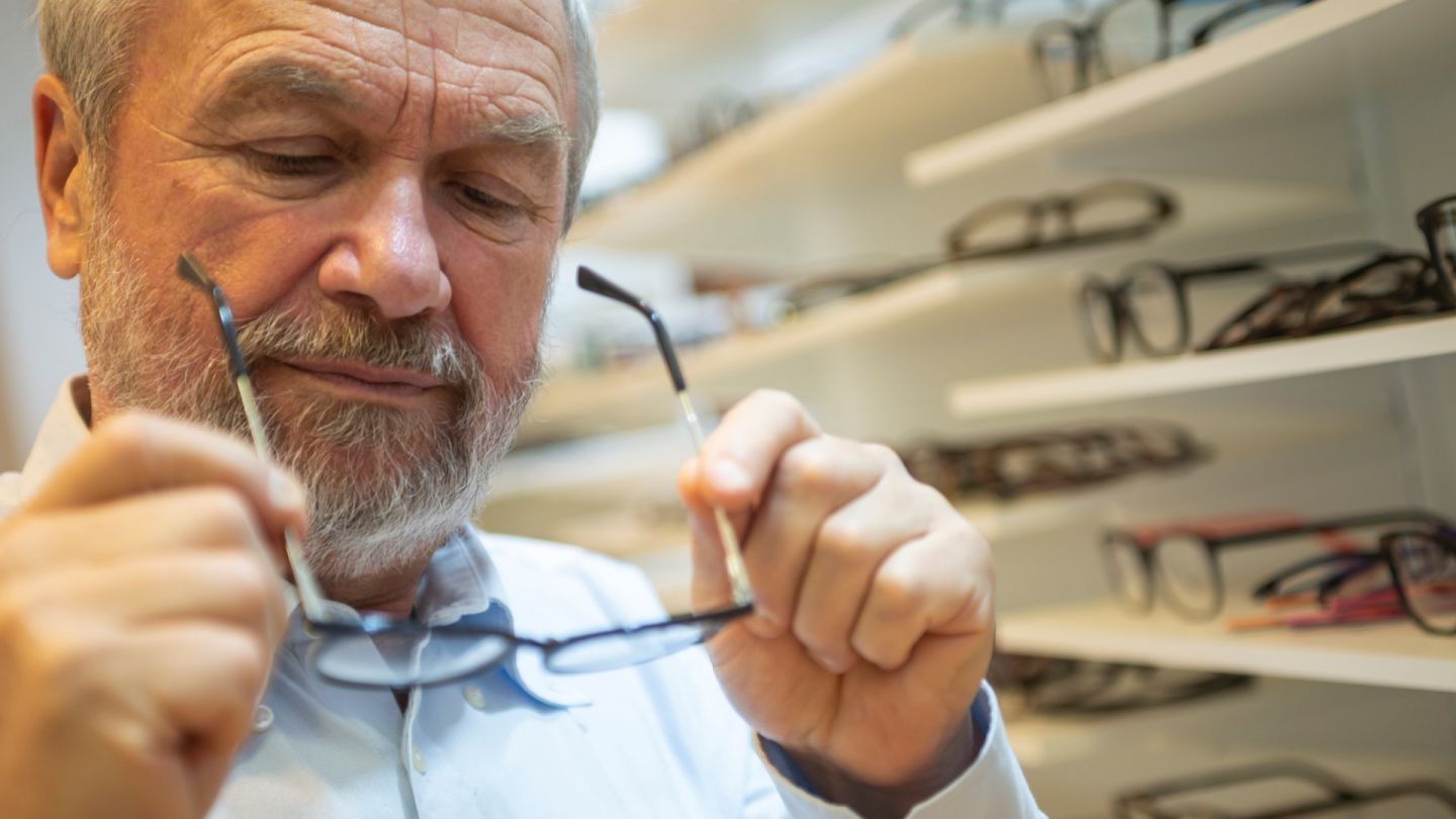 Mann mit Brille (Symbolbild) (Quelle: Getty Images/Koron) Alterssichtigkeit: Ein älterer Mann begutachtet eine Brille, die er in seinen Händen hält.