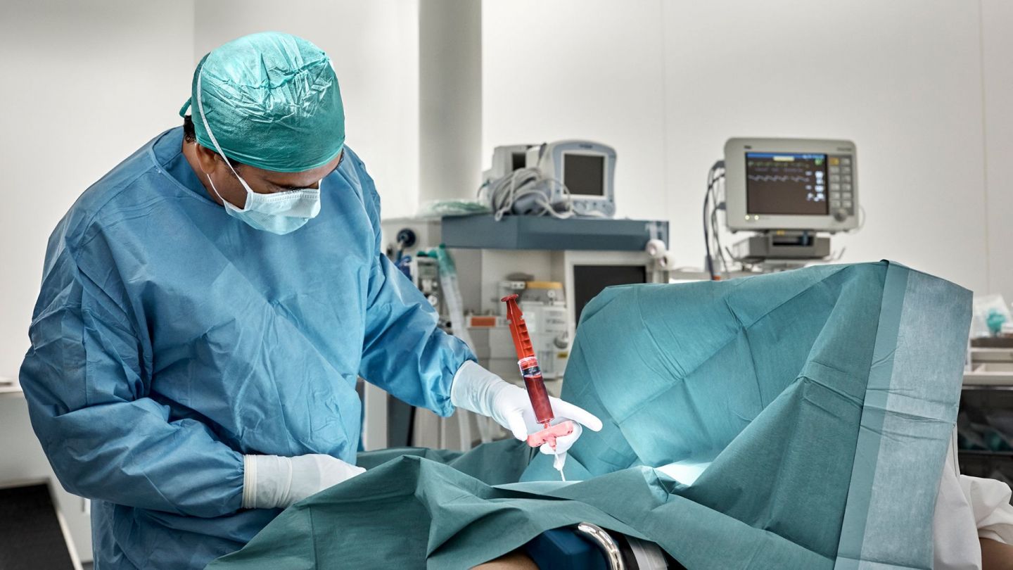 Knochenmarkentnahme (Symbolbild) (Quelle: Getty Images: Morsa Images) Akute myeloische Leukämie bei Erwachsenen: Ein Chirurg steht, in Operationskleidung, in einem Operationssaal. Vor ihm liegt ein abgedeckter Patient auf der Operationsliege. Mit einer Spritze wird ihm Knochenmark entnommen. Der Arzt überwacht den Vorgang.