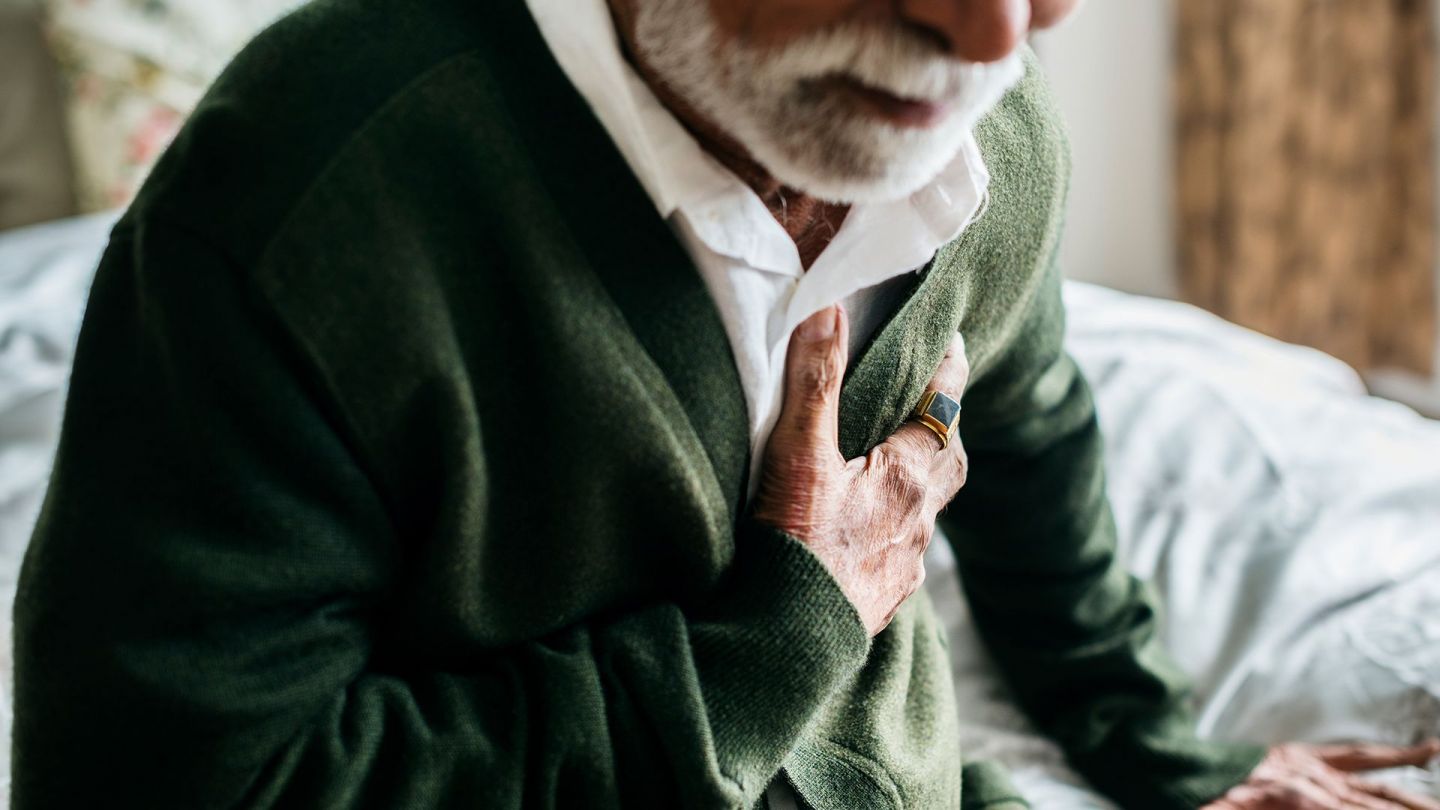 Angina Pectoris - Engegefühl (Quelle: Getty Images/Rawpixel) Angina Pectoris: Ein alter Mann sitzt müde auf einem Bett und fasst sich an den Brustkorb.