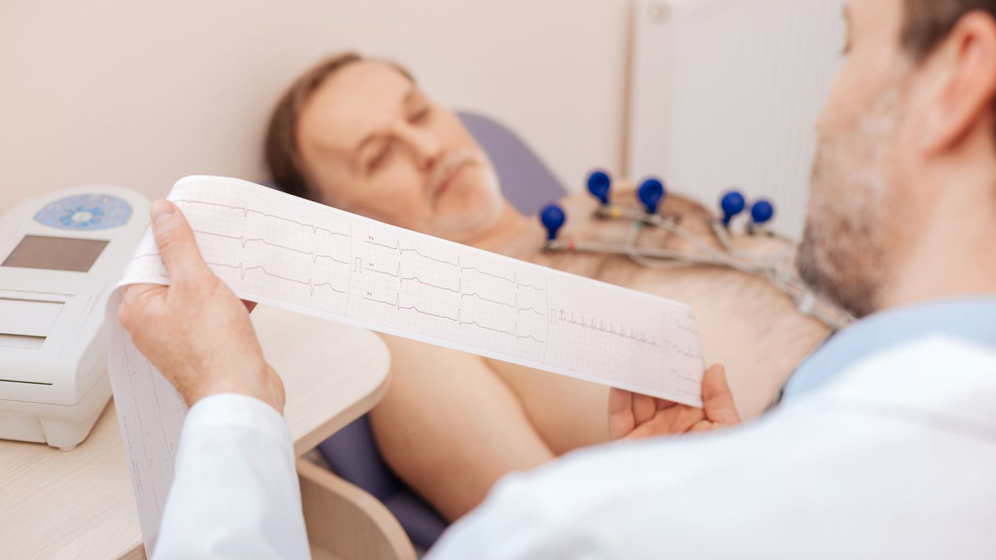 AV-Block-Behandlung (Symbolbild) (Quelle: Getty Images/Zinkevych) AV-Block: Ein Arzt begutachtet die Ergebnisse eines EKG, ein Patient liegt auf einer Behandlungsliege.