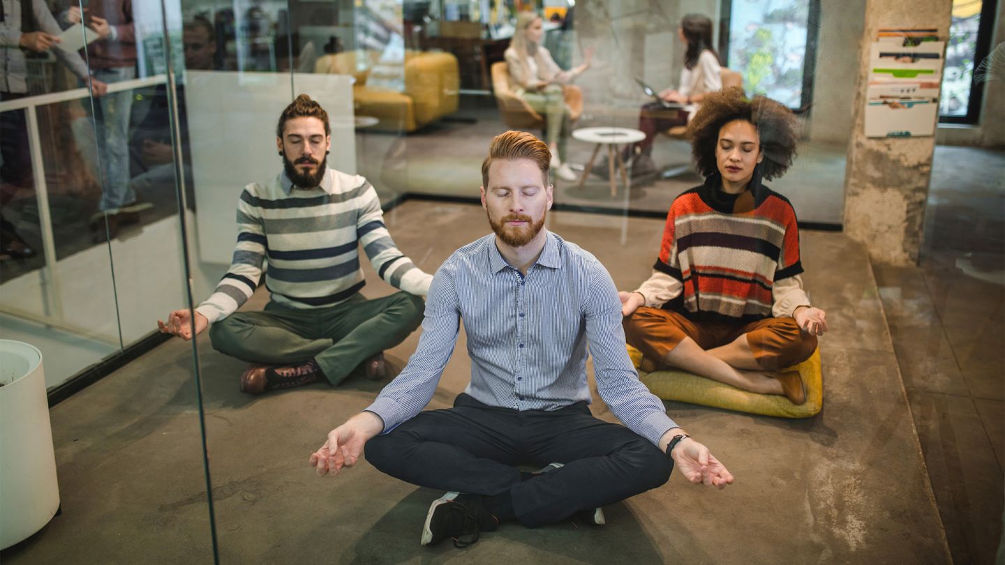 Betriebliche Gesundheitsförderung (Quelle: iStock/skynesher) Betriebliche Gesundheitsförderung: 3 Personen sitzen in einem verglasten Raum in einem Bürogebäude auf dem Boden. Sie sitzen im Schneidersitz und haben ihre Arme auf den Knien abgestützt. Sie scheinen während der Arbeitszeit zu meditieren.