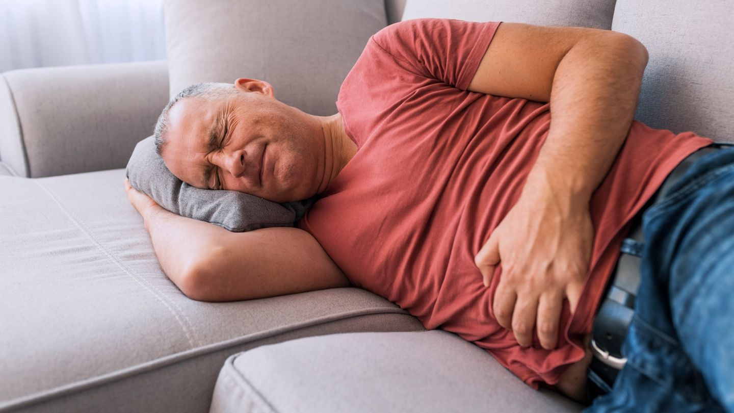 Mann mit Botulismus (Symbolbild) (Quelle: Getty Imagess/ljubaphoto) Botulismus: Ein Mann liegt auf einem Sofa und hält sich mit der linken Hand den Bauch. Seine Augen sind geschlossen und sein Gesicht ist schmerzverzerrt.