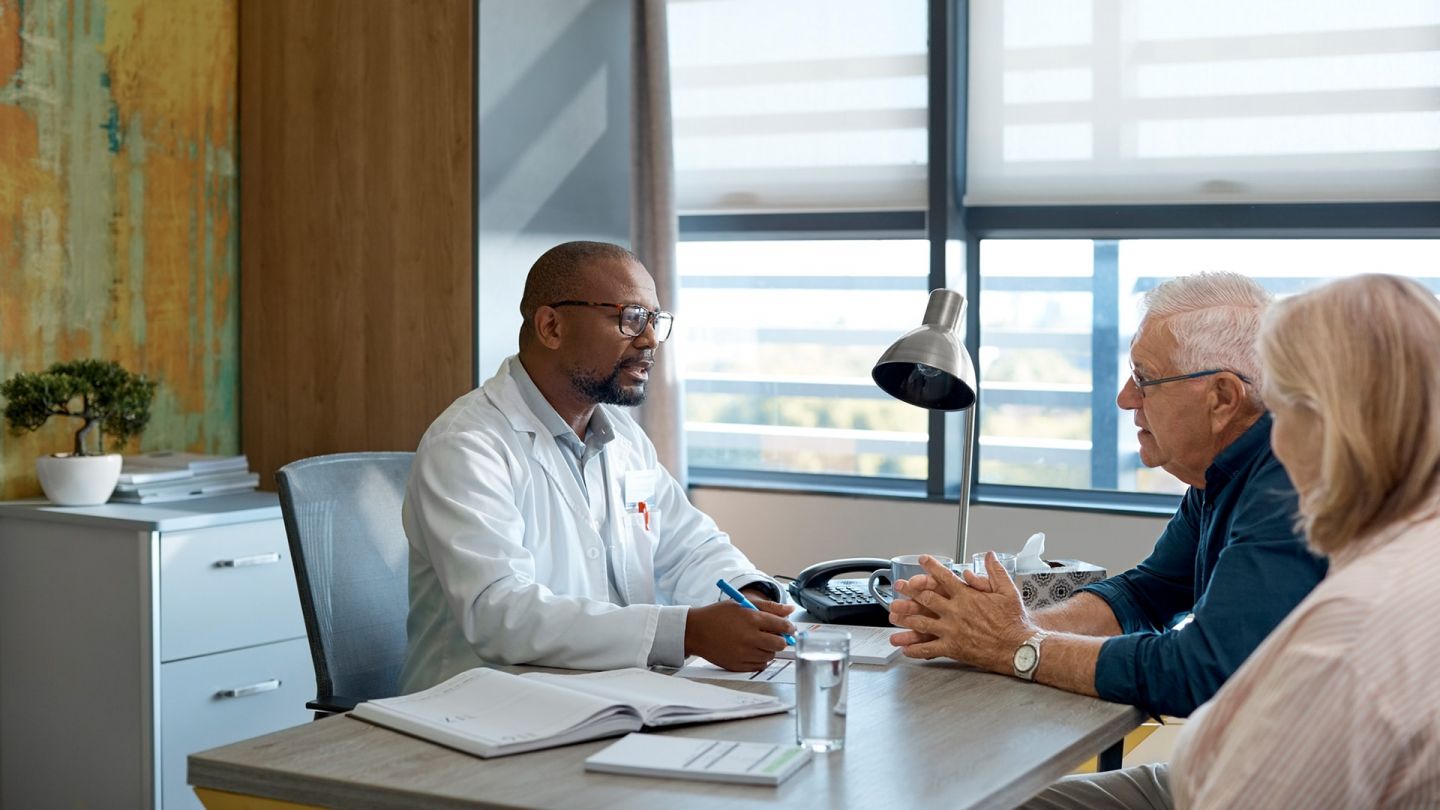 Gespräch mit Patienten (Symbolbild) (Quelle: Getty Images/Morsa Images) Chronische lymphatische Leukämie (CLL): Ein Arzt sitzt an seinem Schreibtisch und führt ein Gespräch mit einem älterern Mann und einer Frau.