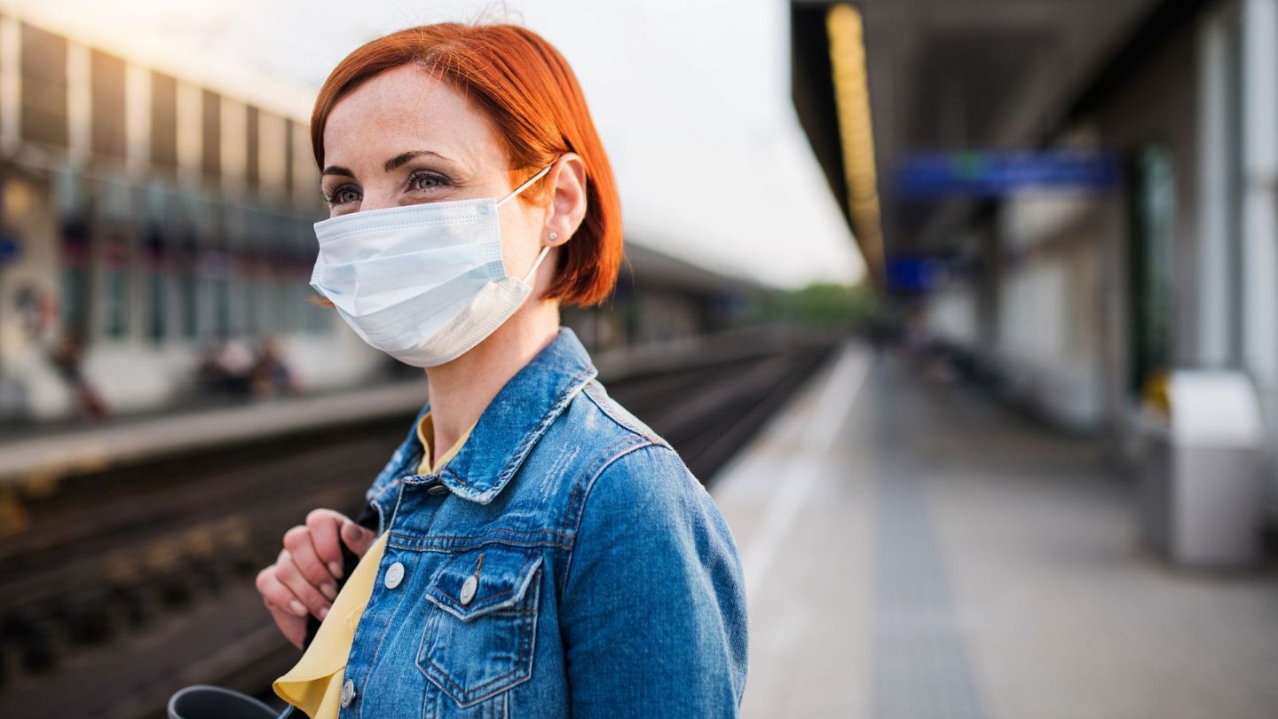 Frau mit Mund-Nasen-Schutzmaske (Quelle: Getty Images/Halfpoint Images) Eine Frau steht an einem verlassenen Bahnsteig und trägt eine Mund-Nasen-Schutzmaske. Mit einer Hand hält sie eine Tasche fest, die an ihrer Schulter liegt. Die Frau schaut in Richtung Bahngleise.