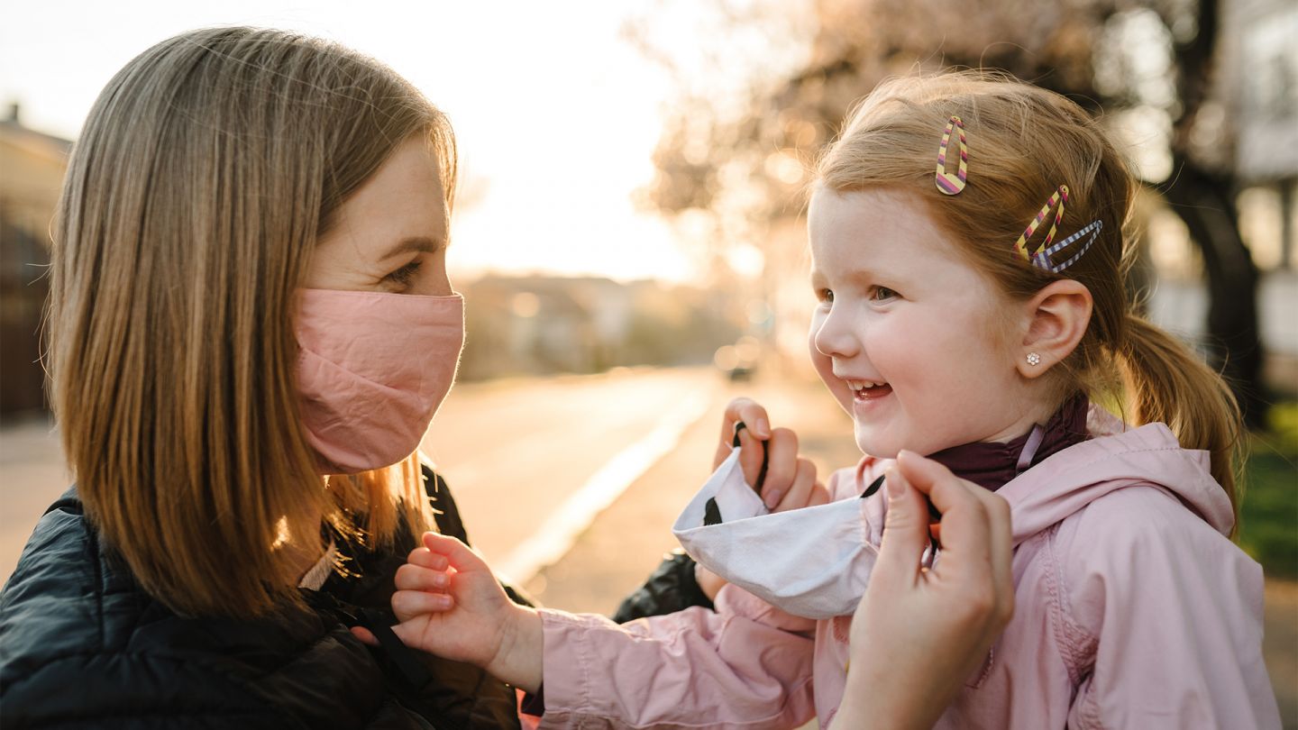 COVID-19 Schutzmaßnahmen (Quelle: iStock/Serhii Sobolevskyi) COVID-19: Eine Mutter kniet draußen auf dem Gehweg vor ihrer kleinen Tochter. Die Mutter trägt eine Mund-Nasen-Bedeckung und setzt ihrer Tochter eine Alltagsmaske auf. Beide wirken entspannt und fröhlich.