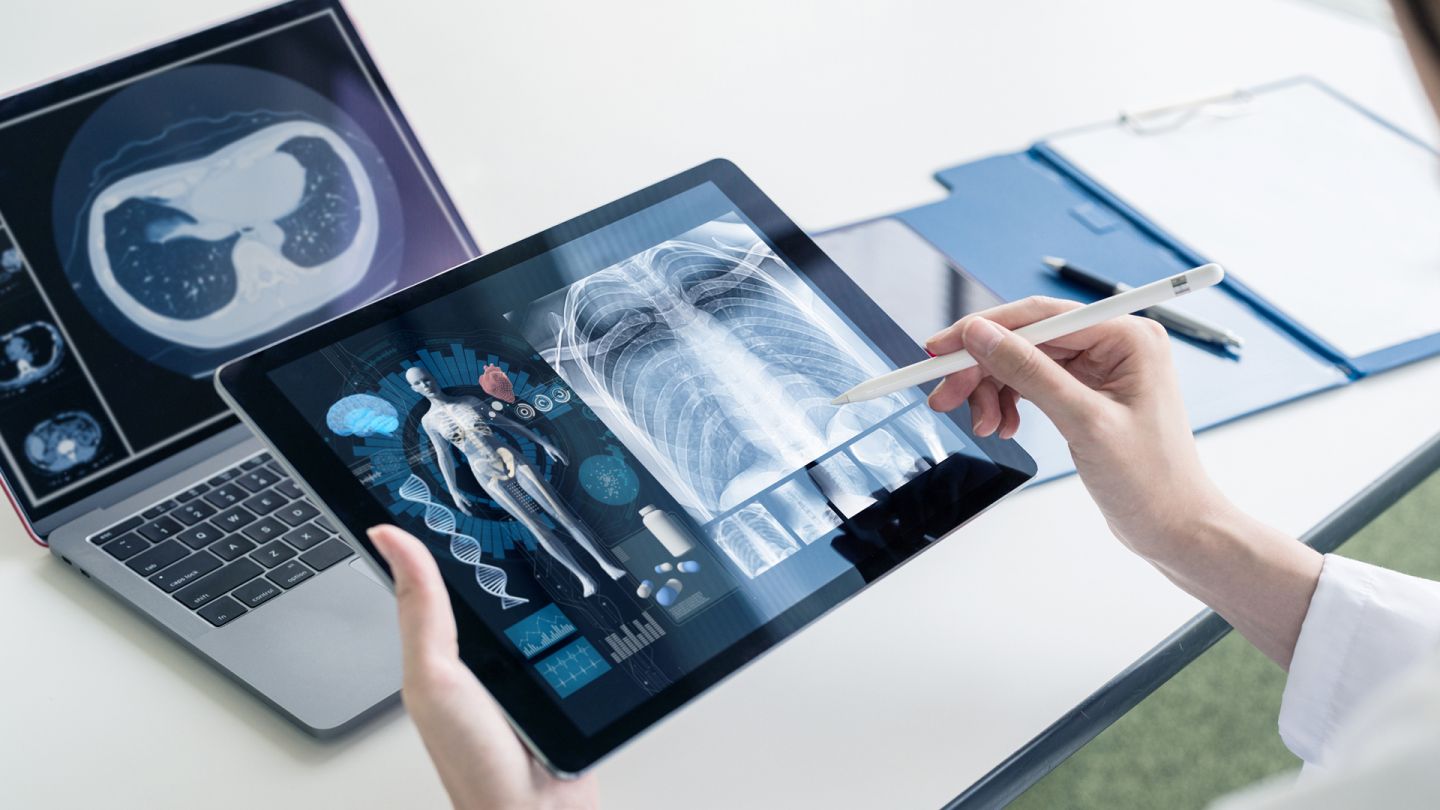 Tablet mit Gesundheitsdaten (Quelle: Getty Images/metamorworks) Notfalldatensatz: Eine Person im weißen Kittel hält ein Tablet in einer Hand – in der anderen Hand einen Stift, der auf den Tablet-Bildschirm zeigt. Dort abgebildet ist das 3D-Modell eines menschlichen Körpers sowie das Röntgenbild eines Rumpfes.