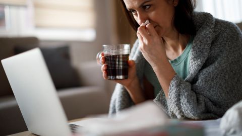 Videosprechstunde bei Erkältung (Quelle: Getty Images/damircudic) Eine Frau schaut auf den Bildschirm eines Laptops, der vor ihr auf einem Tisch steht. Mit einer Hand hält die Frau einen Becher Tee, mit der anderen Hand ein Taschentuch unter ihre Nase. Die Frau ist offenbar erkältet.