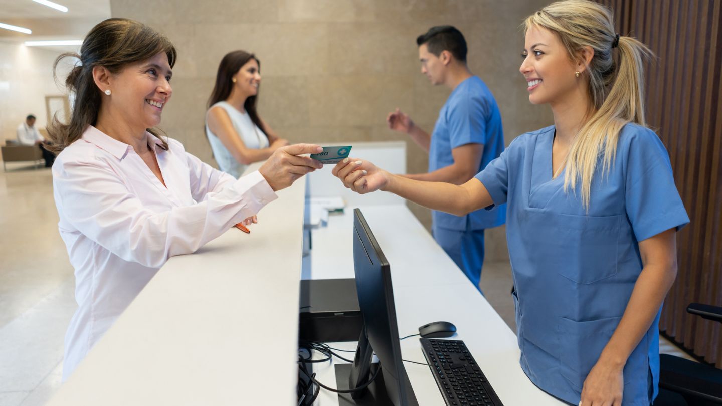 Elektronische Gesundheitskarte (Quelle: Getty Images/Hispanolistic) Elektronische Gesundheitskarte: Eine Patientin und eine Sprechstundenhilfe stehen sich am Empfangsbereich einer Praxis an einem Tisch gegenüber. Die Patientin reicht der Sprechstundenhilfe eine Kunststoffkarte.
