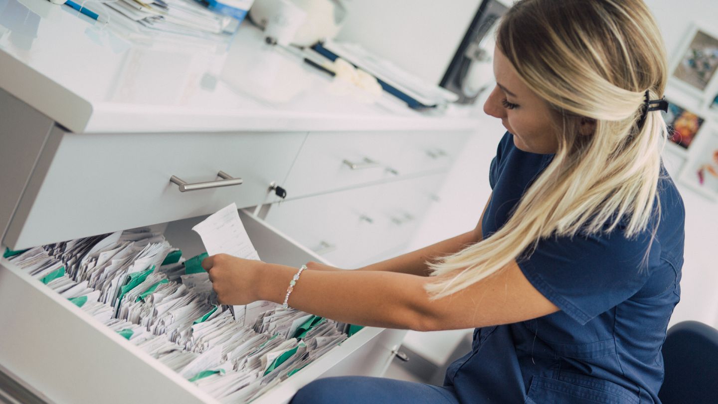 Sprechstundenhilfe und Akten (Quelle: Getty Images/BenAkiba) Elektronische Patientenakte: Eine junge Sprechstundenhilfe sitzt vor der offenen Schublade eines Planschranks, in der einige Dokumente einsortiert sind. Die Frau hält einen Zettel in der Hand und beugt sich nach vorne, um zu lesen, was auf ihm steht.