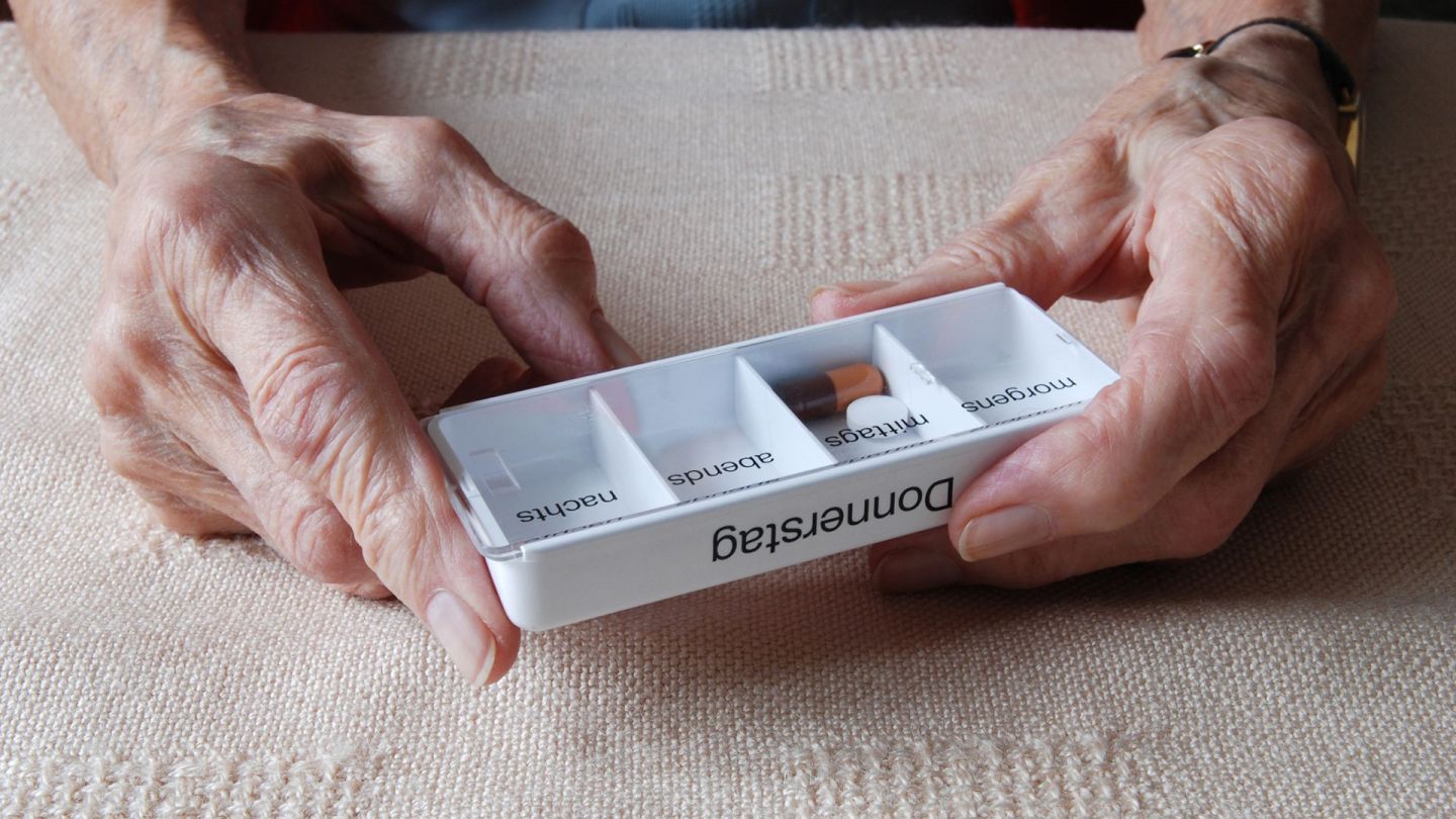 Tablettenbox (Quelle: Getty Images/pixelprof) Elektronischer Medikationsplan (eMP): Eine ältere Frau hält eine Tablettenbox aus Kunststoff in den Händen. Auf der Box steht "Donnerstag". In einem der Fächer liegen eine ovale Tablette und eine Kapsel.