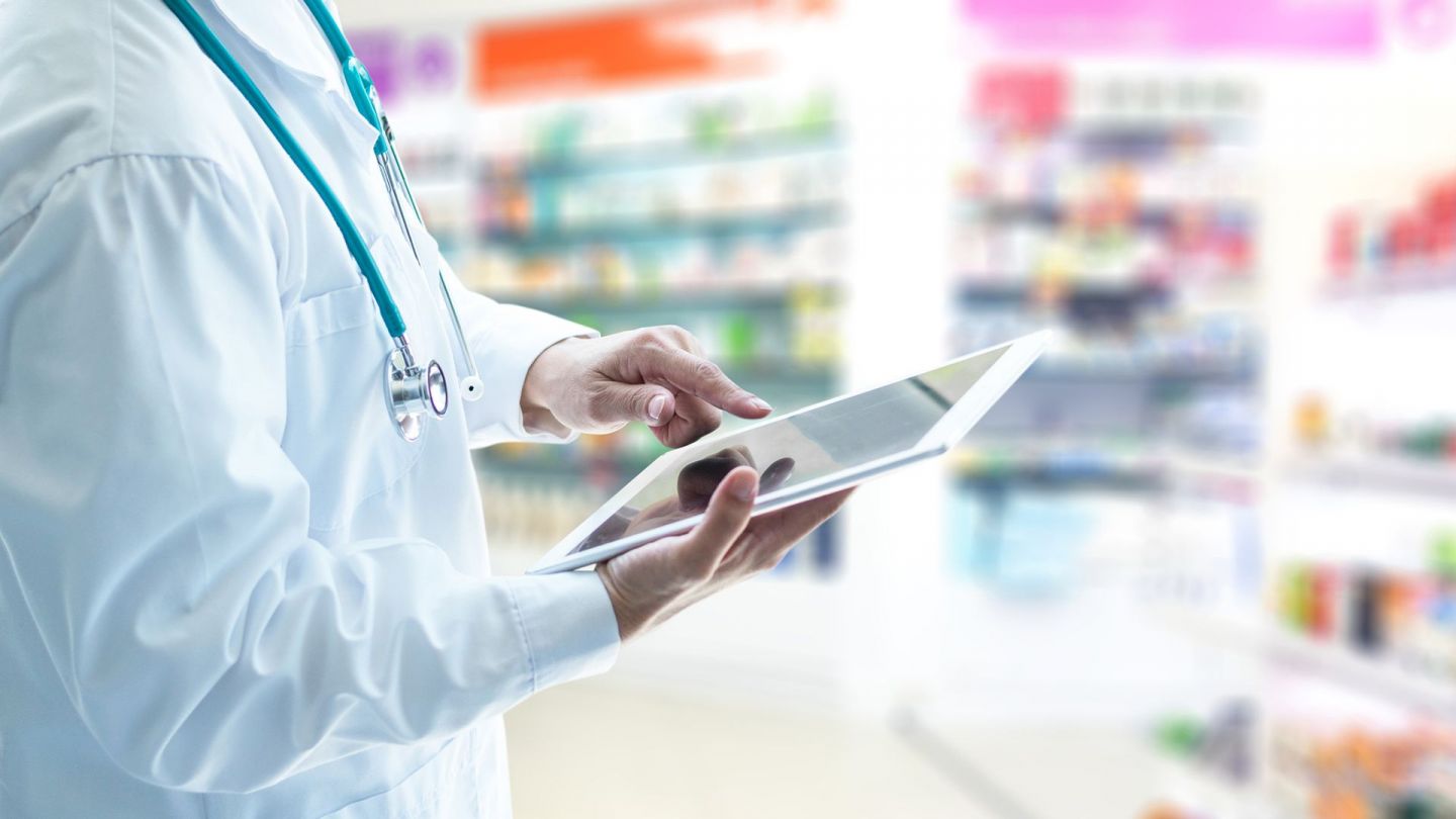E-Rezept in der Apotheke (Quelle: Getty Images/Pornpak Khunatorn) E-Rezept: Ein Mann im weißen Kittel steht in einer Apotheke. In der rechten Hand hält er ein Tablet. Mit dem Zeigefinger der linken Hand deutet der Mann auf den Bildschirm. Im Hintergrund zwei Schränke, in denen Medikamente stehen.