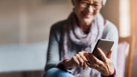 Smartphone mit Gesundheits-App (Quelle: Getty Images/Cecilie_Arcurs) Eine ältere Frau mit Brille sitzt auf einer Couch. Die Frau hält ein Smartphone in der linken Hand, tippt mit der rechten Hand auf den Bildschirm und lächelt.