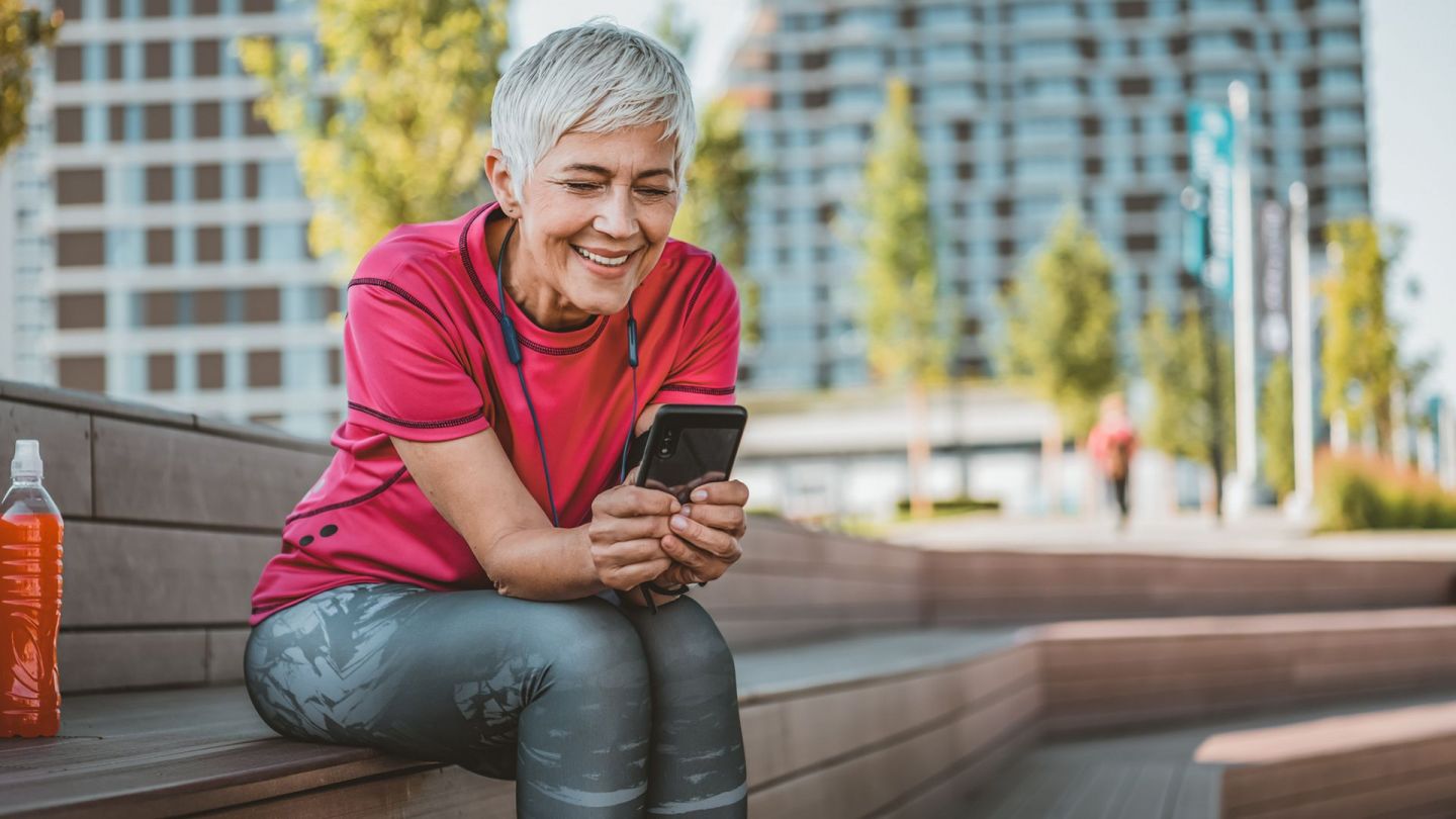 Gesundheits-App für das Smartphone (Quelle: Getty Images/RealPeopleGroup) Digitale Gesundheitsanwendungen (DiGA): Eine Frau sitzt auf einer Holzbank und schaut auf den Bildschirm eines Smartphones, das sie in beiden Händen hält.