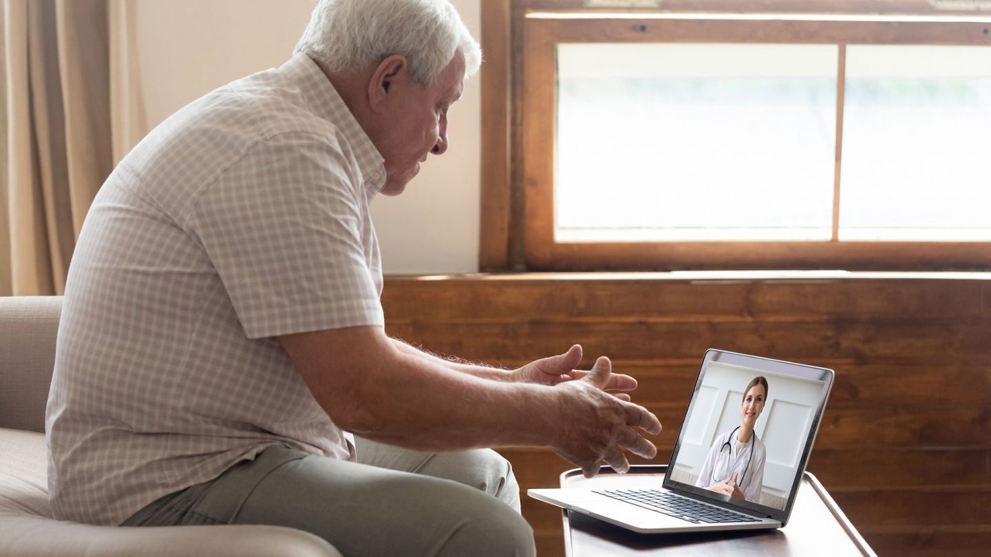 Online-Videosprechstunde mit Ärztin (Quelle: Getty Images/fizkes) Online-Videosprechstunde: Ein älterer Mann sitzt vor einem Laptop, der auf einem Tisch steht. Auf dem Bildschirm sieht er eine jüngere Frau in einem weißen Kittel, die den Mann anschaut.