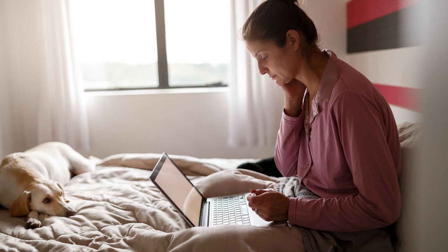 Telemedizin (Symbolbild) (Quelle: Getty Images/Capuski) Telemedizin: Eine Frau sitzt in ihrem Bett und lehnt mit dem Rücken an einem Kissen. Sie hat einen geöffneten Laptop auf ihrem Schoß liegen und schaut auf den Bildschirm. Neben ihr am Rande des Bettes liegt ein Hund, der in Richtung des mobilen PC schaut.
