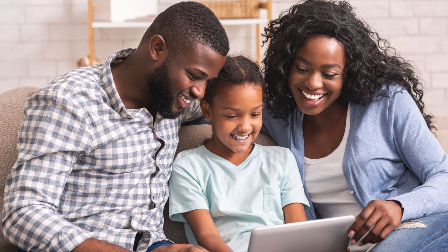 Elektronisches Kinderuntersuchungsheft (Symbolbild) (Quelle: istock/Prostock-Studio) E-Kinderuntersuchungsheft: Mutter, Vater und ein junges Mädchen sitzen gemeinsam auf dem Sofa. Das Mädchen sitzt zwischen den Eltern und hält ein Tablet in der Hand. Alle drei schauen auf den Bildschirm des Geräts.
