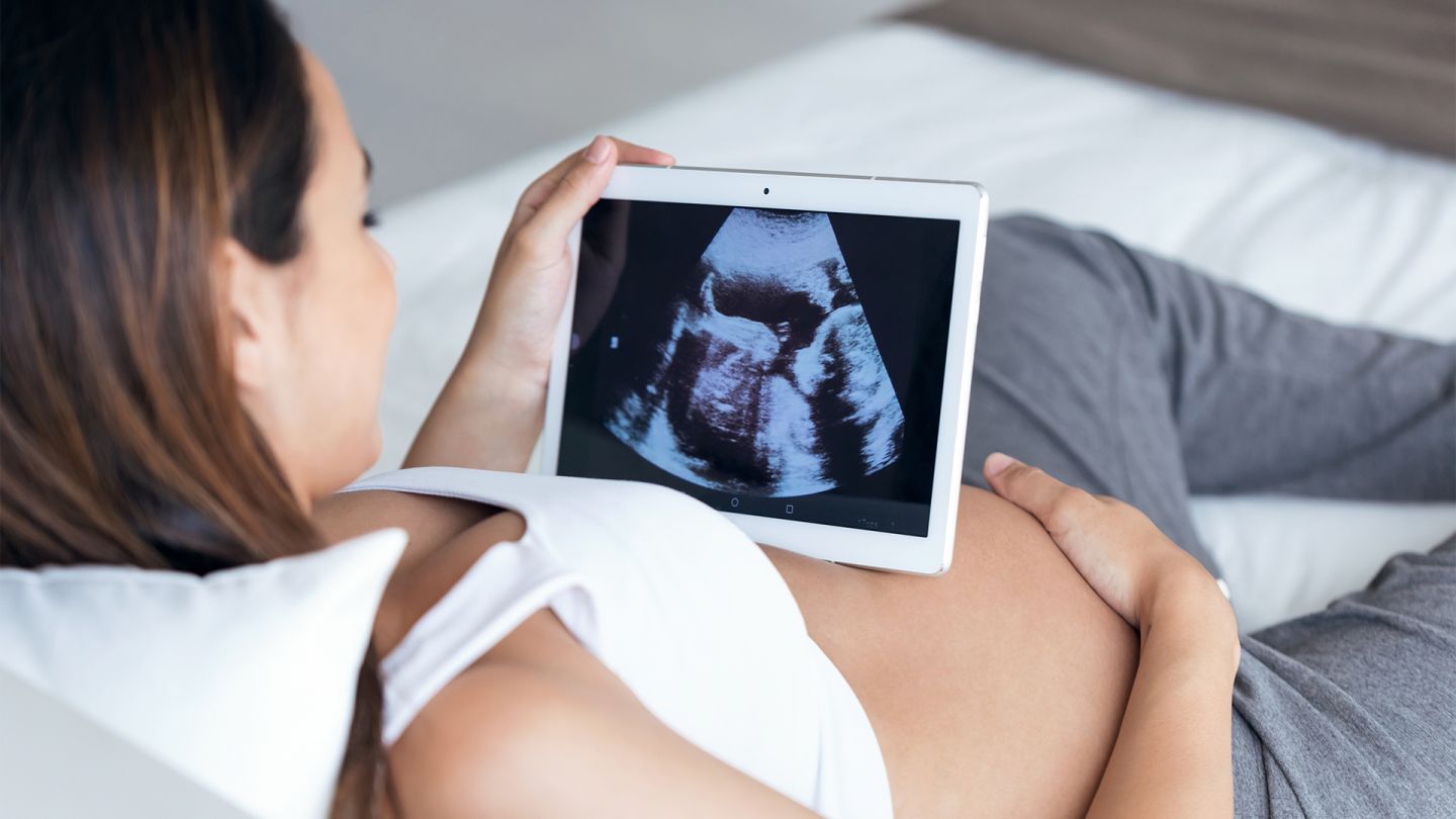 Elektronischer Mutterpass (Quelle: iStock/nensuria) E-Mutterpass: Eine schwangere Frau betrachtet das Ultraschallbild ihres ungeborenen Kindes auf einem Tablet. Das Tablet liegt auf ihrem nackten Bauch, sie wirkt entspannt und zufrieden.