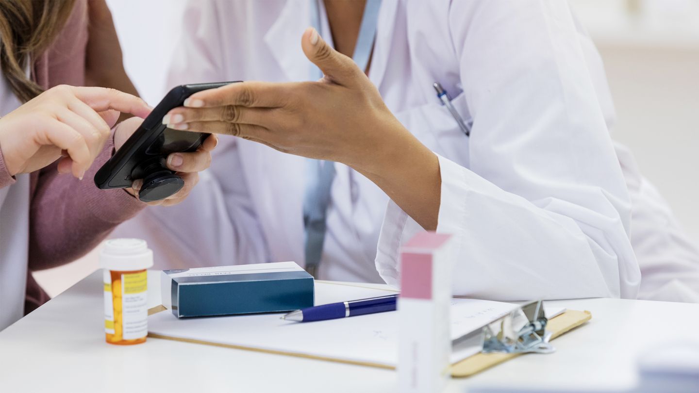 Elektronische Arbeitsunfähigkeitsbescheinigung (Quelle: iStock/SDI Productions) E-Arbeitsunfähigkeitsbescheinigung: Eine Patientin schaut gemeinsam mit einer Ärztin auf ihr Handy. Vor ihnen auf dem Tisch liegen einige Medikamente.