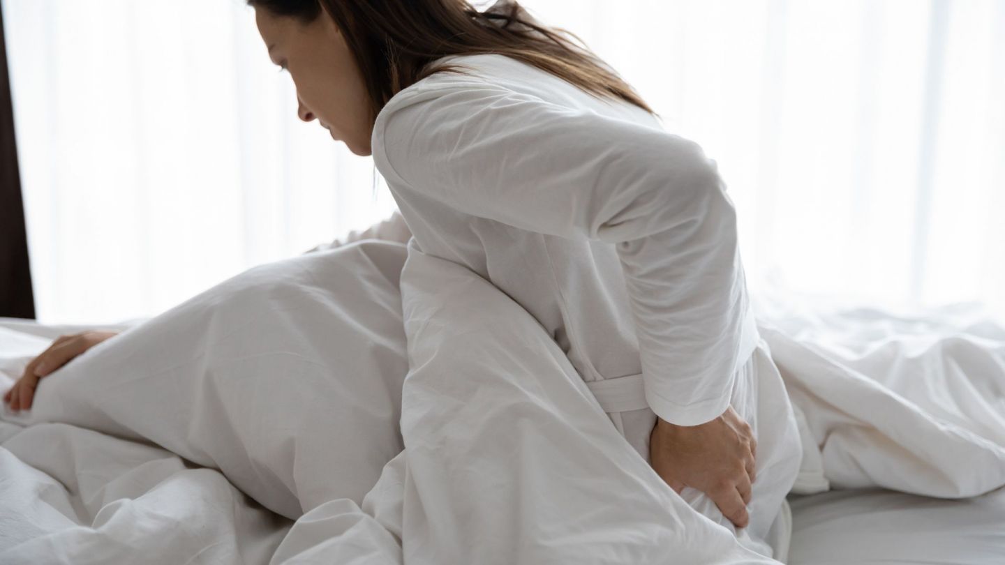 Schmerzen am Rücken (Quelle: Getty Images/fizkes) Fibromyalgie: Eine Frau sitzt leicht nach vorne gebeugt in einem Bett. Mit einer Hand fasst sie sich an den Rücken.