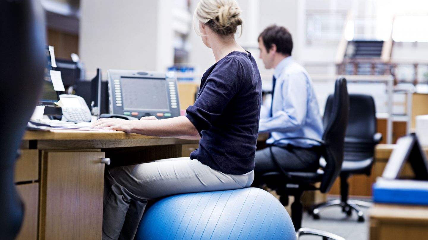Gesundheit am Arbeitsplatz (Quelle: Getty Images/Roger Wight) Gesundheit am Arbeitsplatz: Eine Frau sitzt auf einem Gymnastikball am Schreibtisch und schaut nach rechts zu einem Display.