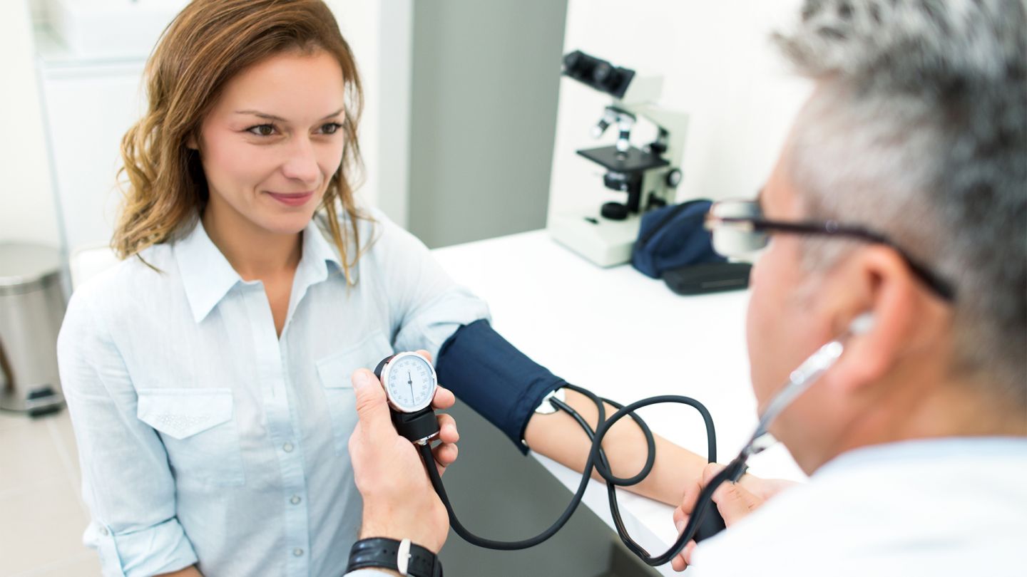 Gesundheits-Check-Up (Quelle: iStock/bluecinema) Gesundheits-Check-Up: Eine Frau sitzt einem Arzt gegenüber. Sie trägt eine Manschette um den Arm, ihr Blutdruck wird gemessen.