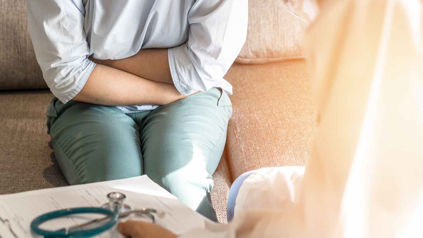 Gonorrhö (Quelle: Getty Images/Chinnapong) Gonorrhö: Eine Frau auf einem Sofa. Sie hält sich mit beiden Händen den Bauch. Sie wirkt angespannt. Ihr gegenüber sitzt ein Arzt.