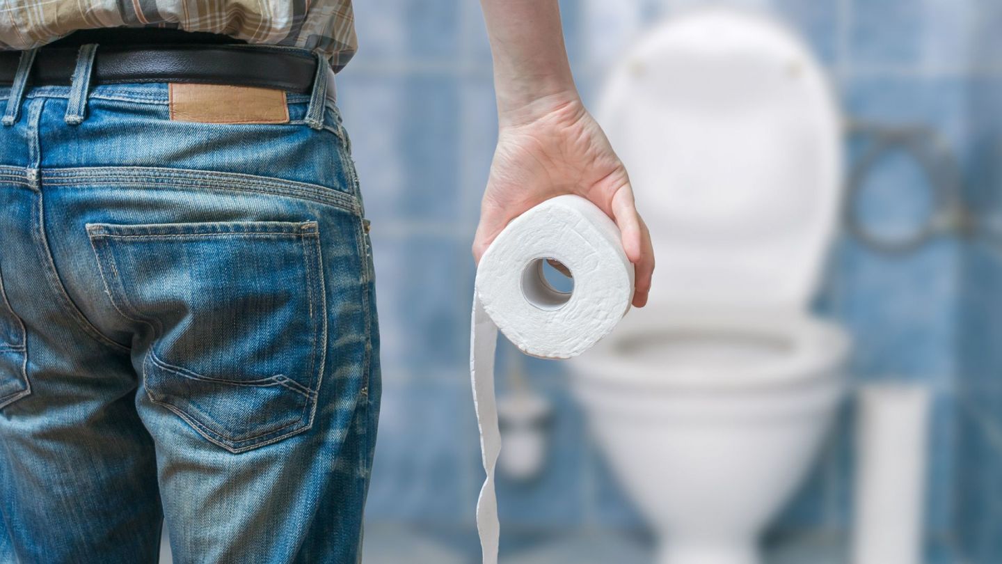 Auf der Toilette (Quelle: Getty Images/vchal) Hämorrhoiden: Ein Mann steht in einem Bad, blickt auf eine Toilette mit geöffneter Klobrille und hält eine Klopapierrolle in der rechten Hand.
