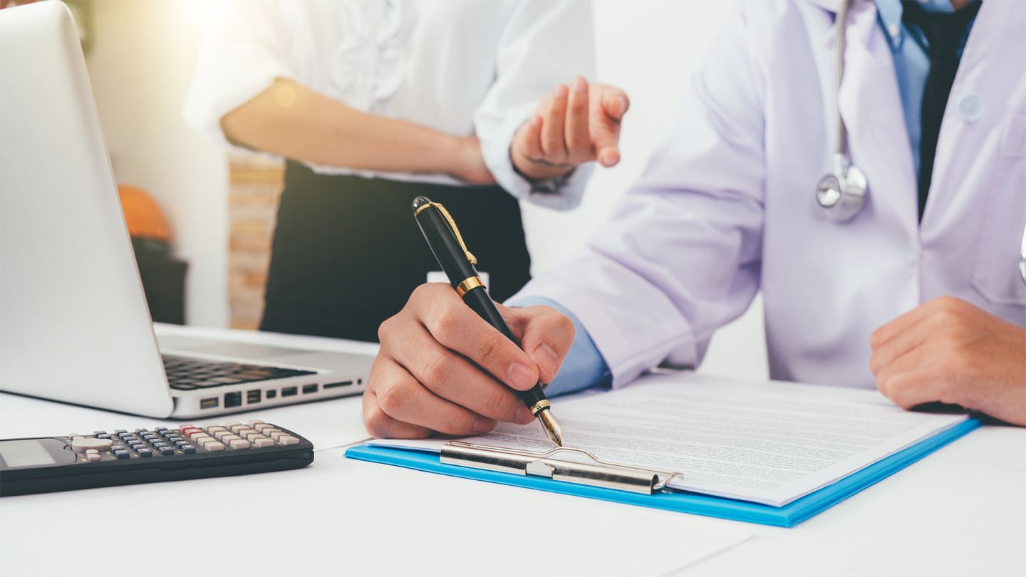 IGeL-Leistungen (Quelle: Getty Images/raybon009) IGeL-Leistungen: Ein Arzt sitzt am Schreibtisch und füllt ein Formular aus. Eine Frau mit weißer Bluse steht hinter ihm. Sie hat den rechten Arm vor dem Bauch verschränkt und mit dem anderen Arm macht sie eine Handbewegung in Richtung des Arztes.