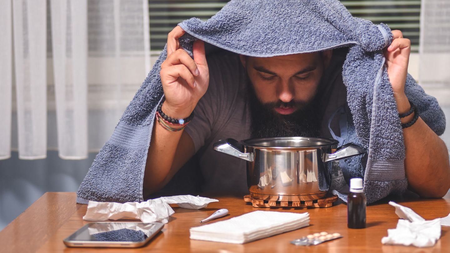 Erkältung (Symbolbild) (Quelle: Getty Images/Ika84) Nasennebenhöhlenentzündung: Ein Mann sitzt an einem Tisch, auf dem Papiertaschentücher, ein Fieberthermometer, Tabletten, eine Flasche Nasentropfen und ein Topf stehen. Der Mann hält ein Handtuch über seinen Kopf, beugt sich über den Topf und inhaliert.
