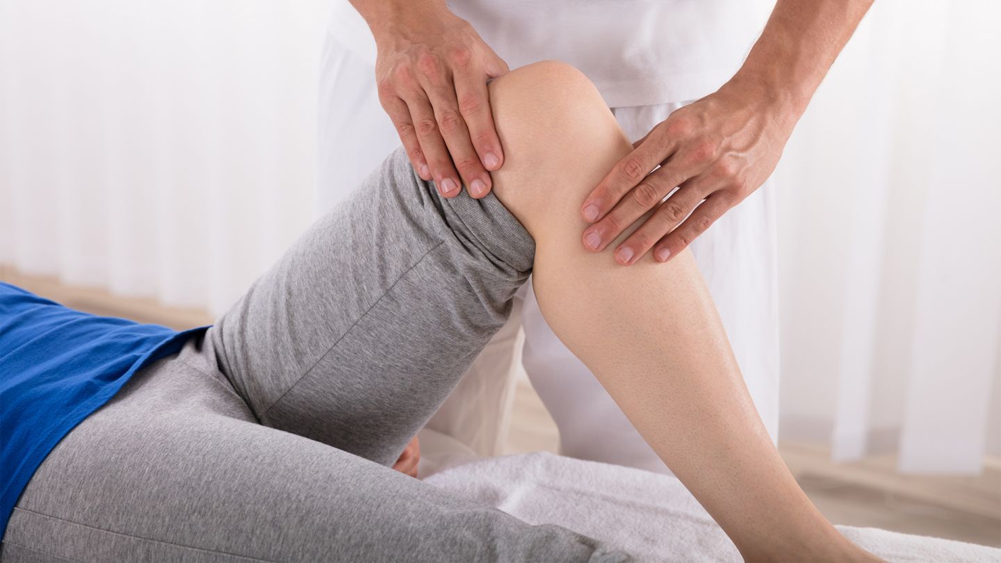 Patellaluxation (Quelle: iStock/AndreyPopov) Patellaluxation: Eine junge Frau liegt auf einer medizinischen Liege. Ein Physiotherapeut tastet mit beiden Händen ihr gebeugtes Knie ab.