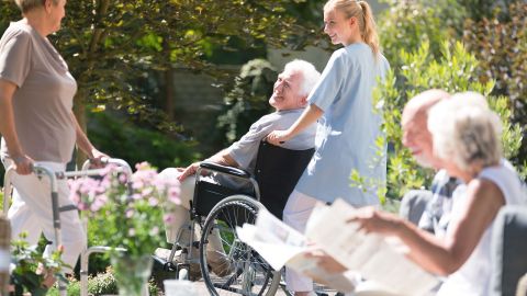 Senioren und Pflegerin im Garten (Quelle: Getty Images/KatarzynaBialasiewicz) Vollstationäre Kurzzeitpflege: Eine Pflegerin schiebt einen älteren Mann, der im Rollstuhl sitzt. An ihnen läuft eine ältere Frau mit Gehhilfe vorbei. In der Nähe sitzen ein Mann und eine Frau, die jeweils eine Zeitung in der Hand halten und lesen.