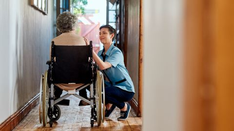 Pflege zu Hause Definition (Symbolbild) (Quelle: Getty Images/Dean Mitchell) Pflege von zu Hause: Eine alte Dame im Rollstuhl steht im Hausflur auf dem Weg nach draußen. Ihre häusliche Pflegerin spricht mit ihr und hockt dabei neben der Dame, um mit ihr auf Augenhöhe zu sein.