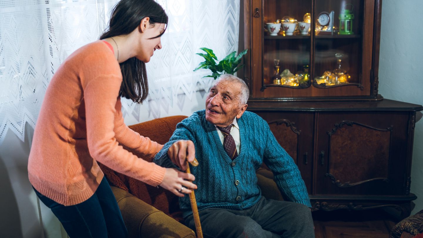 Pflege zu Hause (Symbolbild) (Quelle: Getty Images/urbazon) Ein alter Herr möchte aus seinem Wohnzimmersessel aufstehen, er hat einen Gehstock zur Hilfe. Eine junge Frau hilft ihm bei dem Aufstehen und greift ihm unter den Arm als Stütze.