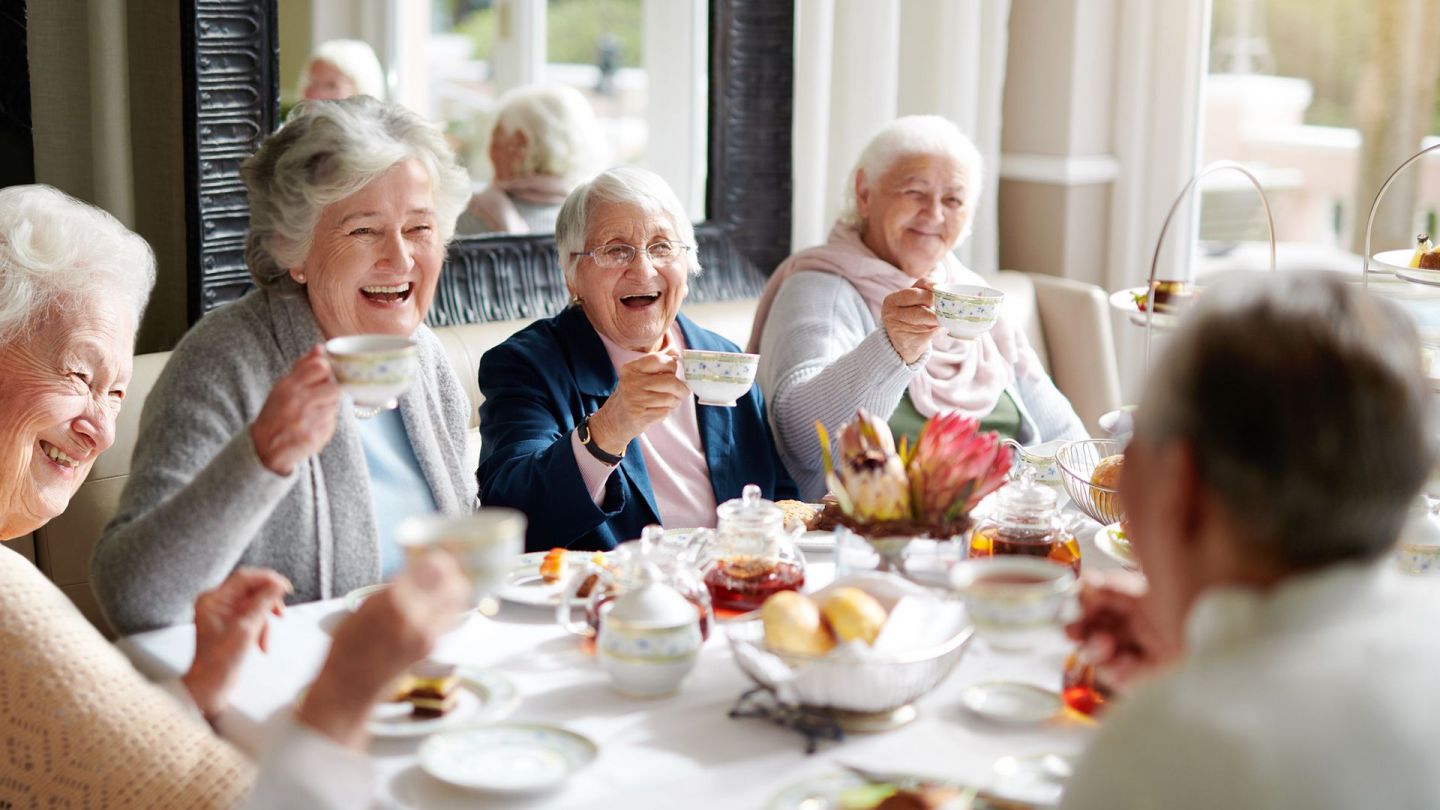 Frühstück in einer Pflege-WG (Quelle: Getty Images/Cecilie_Arcurs) Pflege-WGs – Leben in ambulant betreuten Wohngruppen: Fünf Seniorinnen sitzen an einem Esstisch, auf dem unter anderem Schälchen mit Brötchen und Marmelade stehen. Alle Frauen haben eine Tasse in der Hand, heben sie an und prosten sich zu.