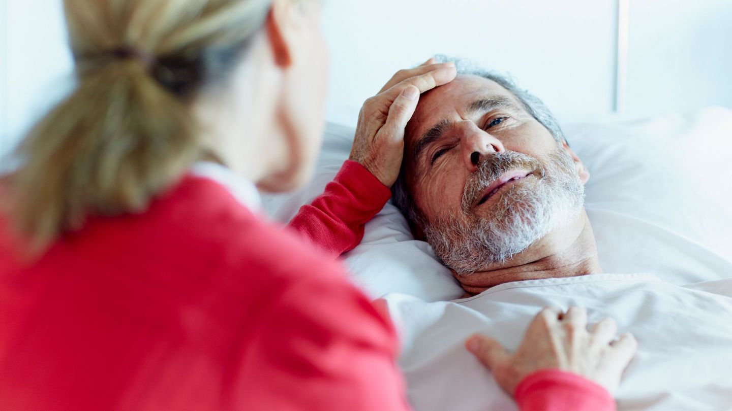 Kranker Mann (Symbolbild) (Quelle: Getty Images/Morsa Images) Leistungen für schwer kranke und sterbende Menschen: Ein Mann mittleren Alters liegt in einem Krankenbett. Er schaut leicht zur Seite an den Bettrand. Dort sitzt eine Frau. Mit ihrer linken Hand berührt sie die Stirn des Mannes.