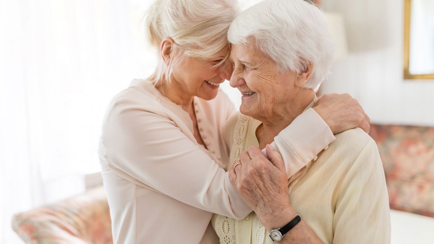 Mutter und Tochter (Quelle: Getty Images/Piksel) Pflegeversicherung – Hilfe für pflegende Angehörige: Mutter und Tochter stehen sich gegenüber und lachen. Die Tochter legt ihren Arm um die rechte Schulter der Seniorin. Sie wiederrum hält sich mit einer Hand am Arm der jüngeren Frau fest.