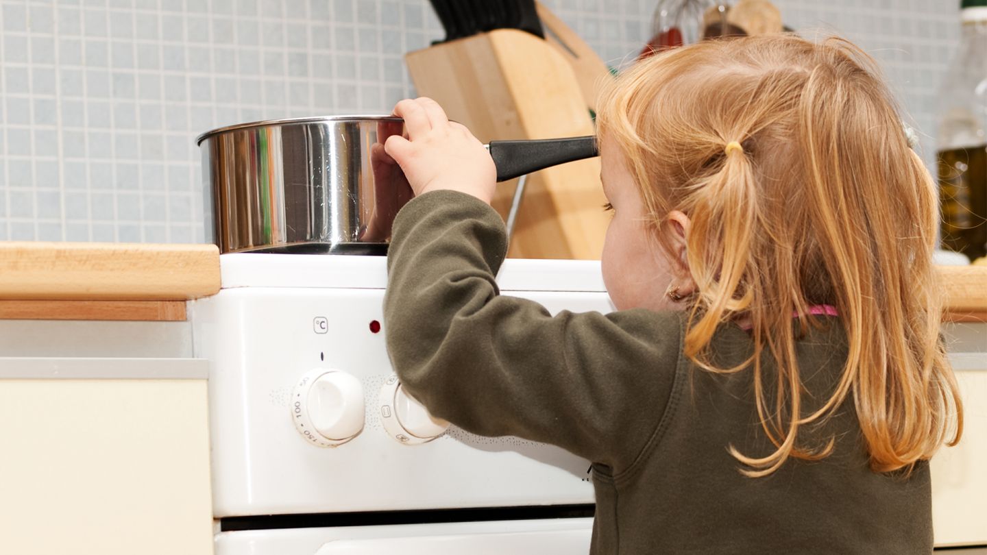 Prävention von Kinderunfällen (Quelle: Getty Images/Libortom) Ein kleines Mädchen steht vor einem Küchenherd auf dem ein Topf mit kochendem Inhalt steht und greift an den Topfrand, um in den Topf rein zu schauen.