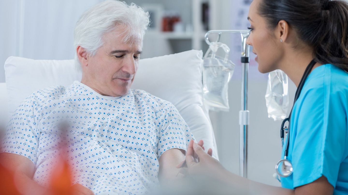 Patient und Krankenschwester (Symbolbild) (Quelle: Getty Images/SDI Productions) Prostatakrebs: Ein älterer Mann sitzt in einem Krankenhausbett. Er schaut nach unten, ihm gegenüber sitzt eine Krankenschwester. Sie schaut ihn an und entnimmt ihm Blut.