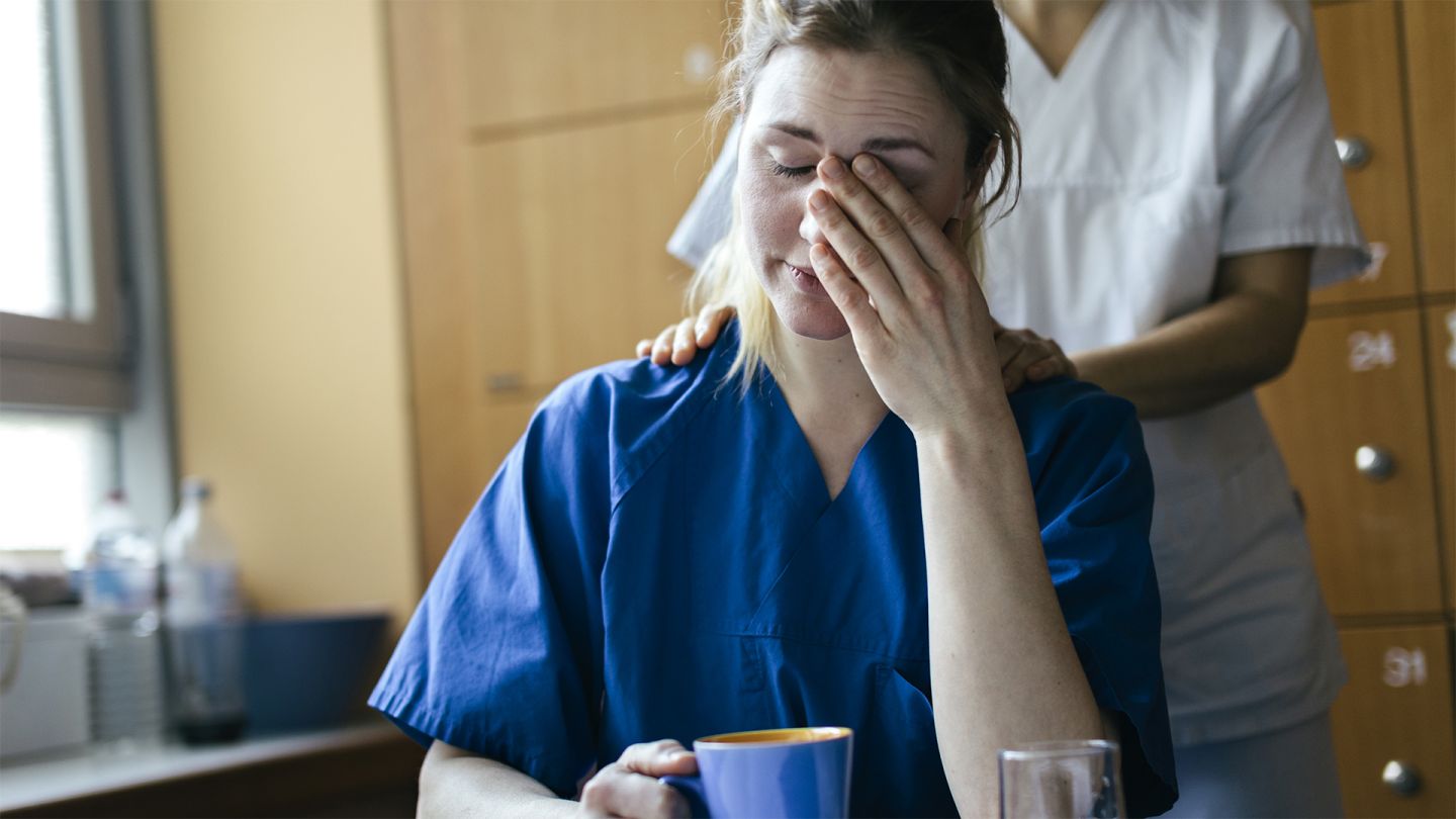 Psychische Gesundheit am Arbeitsplatz (Symbolbild) (Quelle: Getty Images/Hinterhaus Productions) Psychische Gesundheit am Arbeitsplatz: Eine Krankenschwester sitzt mit einer Tasse an einem Tisch. Mit dem linken Ellenbogen stützt sie sich auf und reibt mit ihrer Hand über ihr Gesicht. Eine Kollegin legt ihr von hinten tröstend die Hände auf die Schul