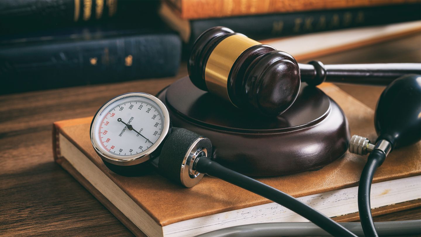 Rechte für Patienten (Getty Images/Rawf8) Rechte für Patienten: Ein mechanisches Blutdruckmessgerät liegt zusammen mit einem Richterhammer auf einem großen Buch, welches auf einem Massivholztisch liegt. Im Hintergrund liegen Bücher.