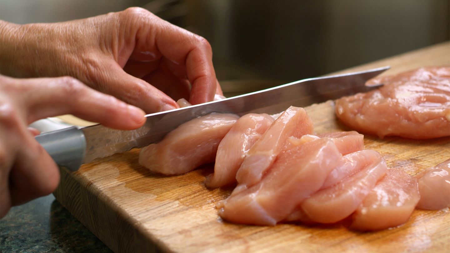 Salmonellen (Quelle: iStock/stephanie phillips) Die Hände einer Frau schneiden Hühnchenfleisch in Scheiben. Das Fleisch liegt auf einem hölzernen Schneidebrett.