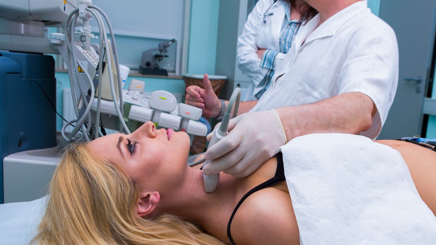 Ultraschalluntersuchung der Schilddrüse (Quelle: Getty Images/choja) Schilddrüsenüberfunktion: Eine junge Frau liegt auf der Behandlungsliege in einer Praxis. Ein Arzt, der einen Medizinhandschuh trägt, hält der Frau ein Messgerät an den Kehlkopf. Der Arzt untersucht ihre Schilddrüse per Ultraschall.