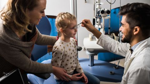 Mädchen beim Augenarzt (Quelle: Getty Images/skynesher) Amblyopie: Ein Mädchen sitzt auf einem Behandlungsstuhl in einer Praxis. Vor ihr kniet ein Augenarzt. Mit einer Hand hält er eine Brille vor die Augen des Mädchens. Eine Frau steht daneben und stützt den Oberkörper des Kindes.