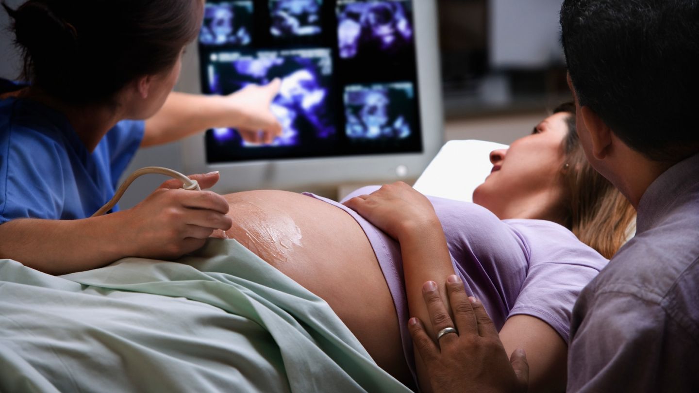 Schwangerschaftsvorsorge (Quelle: Getty Images/Jose Luis Pelaez Inc) Schwangerschaftsvorsorge: Eine Schwangere wird von einer Frauenärztin mit einem Ultraschallgerät untersucht. Ihr Mann sitzt neben ihr und legt behutsam seine Hand auf ihren Arm. Beide schauen auf den Monitor, auf dem die Ärztin auf die Bilder zeigt.
