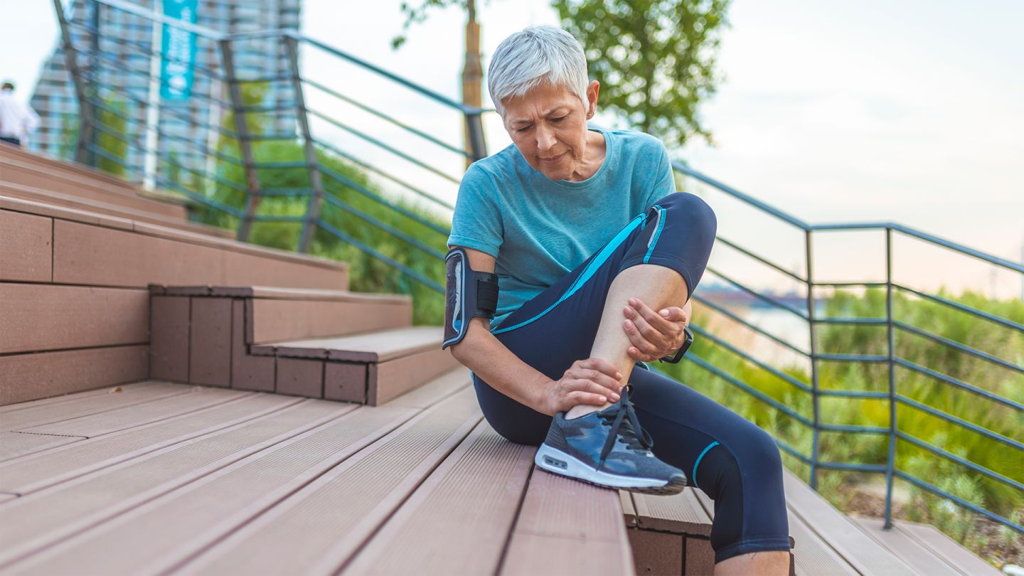 Sehnenreizung (Quelle: iStock/dragana991) Sehnenreizung: Eine ältere Frau in Sportkleidung sitzt im Freien auf einer Treppe. Sie hält sich den Knöchel, ihr Gesicht ist schmerzverzerrt.