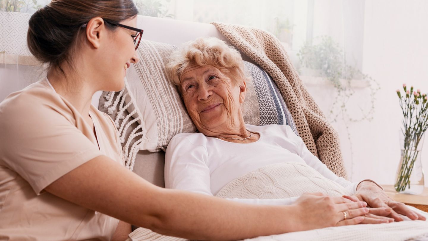 Ältere Frau mit Krankenschwester (Symbolbild) (Quelle: Getty Images/KatarzynaBialasiewicz) Seltene Krebsarten: Eine ältere Frau liegt in einem Bett. Neben ihr sitzt eine Krankenschwester und hält ihre Hand. Die beiden lächen sich an.