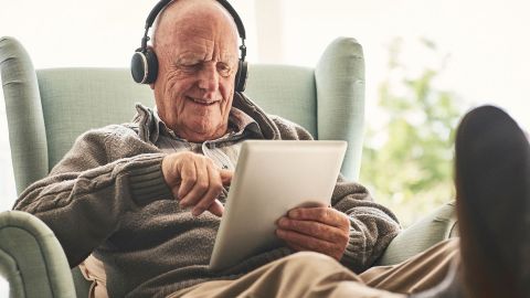 Informationen für Patienten im Internet (Quelle: Getty Images/Dean Mitchell) Ein älterer Mann sitzt mit ausgestreckten Beinen auf einem Sessel. Er trägt Kopfhörer und schaut lächelnd auf den Bildschirm eines Laptops, den er in seiner linken Hand hält. Mit dem Zeigefinger der anderen Hand berührt der Mann den Bildschirm.