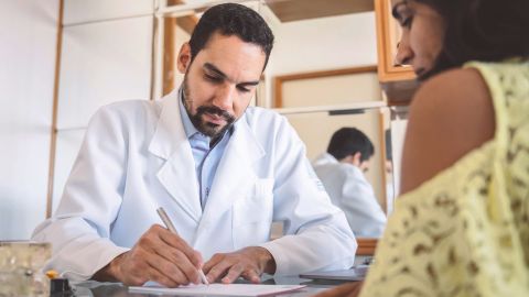 Arztbeleg für Patientin (Quelle: Getty Images/golero) Ein Arzt und eine Frau sitzen gemeinsam an einem Tisch. Der Arzt hält einen Kugelschreiber in der Hand und schreibt auf ein Blatt Papier. Er stellt einer Patientin eine Quittung aus.