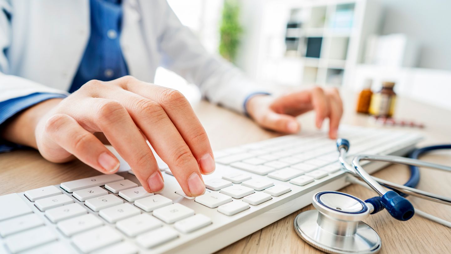 Recherche (Quelle: Getty Images/seb_ra) Zweitmeinung: Eine Ärztin sitzt an einem Schreibtisch und tippt mit den Fingern beider Hände auf eine Computertastatur. Neben der Tastatur liegt ein Stethoskop.