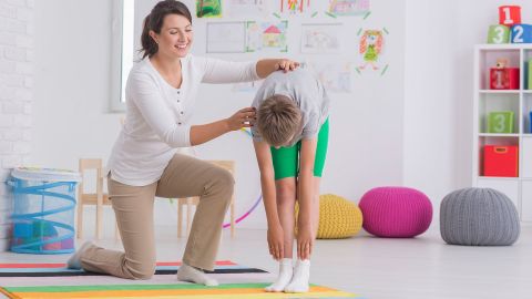 Physiotherapie bei Skoliose (Quelle: Getty Images/KatarzynaBialasiewicz) Eine lächelnde Physiotherapeutin stützt einen Jungen, der sich stehend nach vorne beugt und versucht, mit den Händen seine Füße zu berühren.